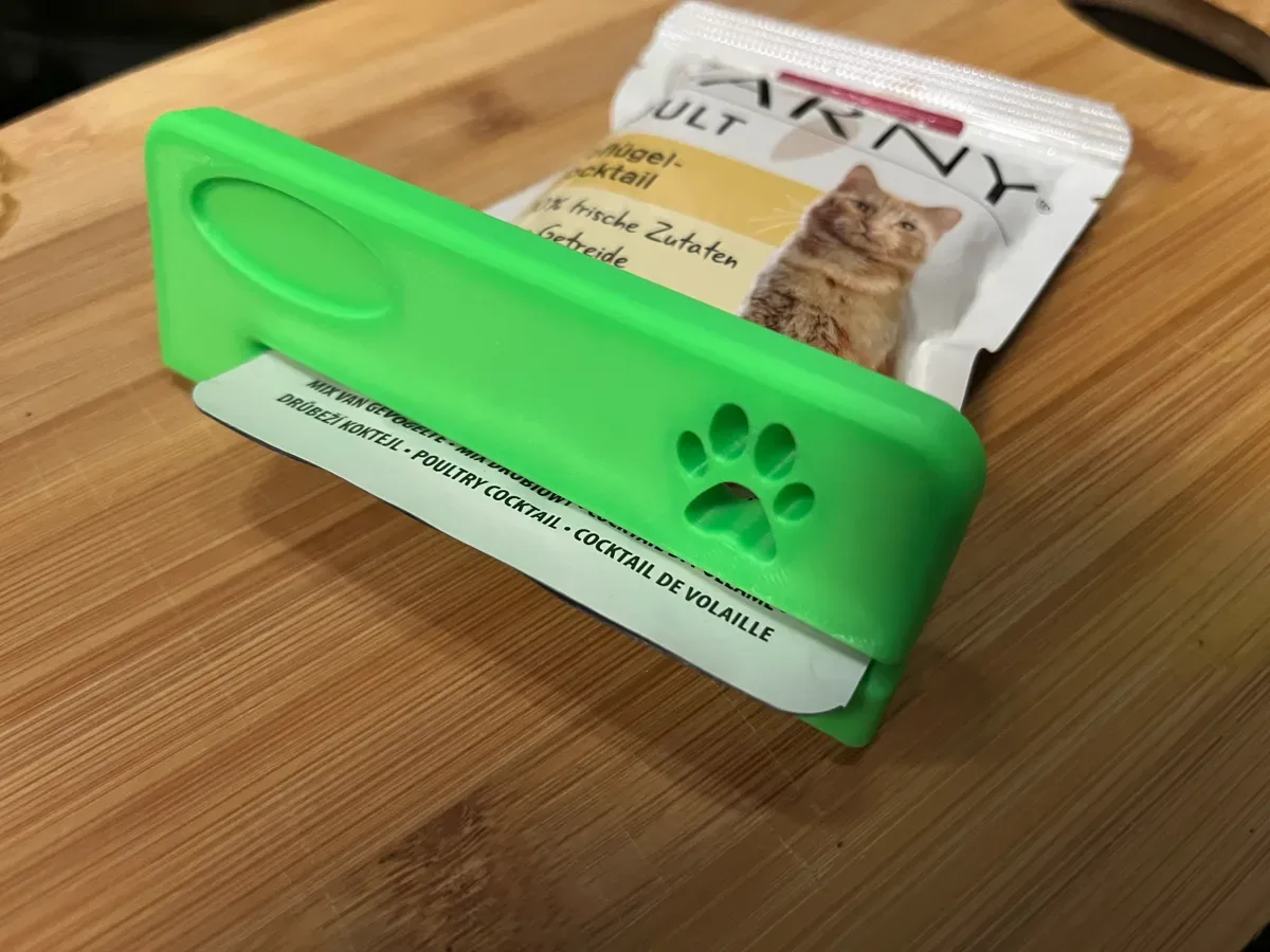 Der grüne Abstreifer mit Pfotenabdruck steckt auf einem Katzenfutterbeutel, der Beutel liegt auf einem Holzbrett