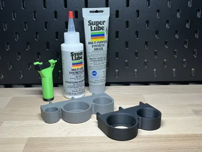 Two grey holders on the wooden surface in front of the black pegboard, behind them the Super Lube and Synco Lube bottles and a green grease gun