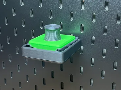 The grey holder hangs on the pegboard with the green build plate cleaner and its brush standing in it