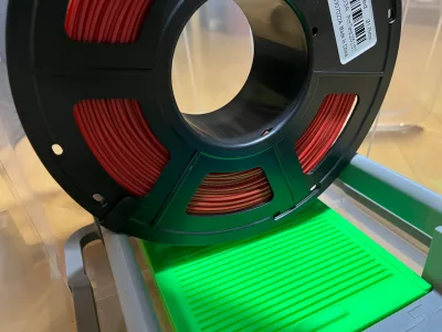 A black spool of red filament sits in the holder, below it the green box with its ribbed lid