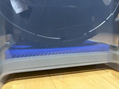 Side view into the clear crate: the blue box lies flat under a filament spool, its diagonally printed vents visible