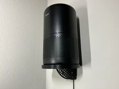 The black air purifier stands on the round wall shelf against a white wall, the cable leaving the channel at the bottom